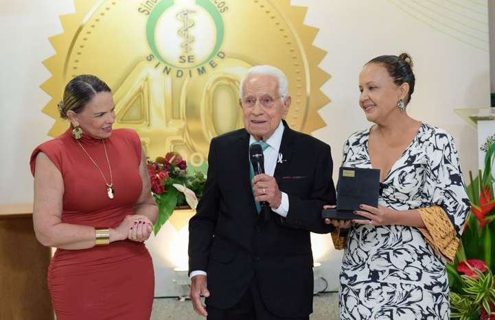 Dr. Raulino Galrão Lima discursando ao microfone ao lado de duas mulheres