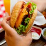 Mão segurando hambúrguer com refrigerante ao fundo