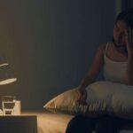 Mulher sentada na cama à noite com a mão na cabeça, sem conseguir dormir